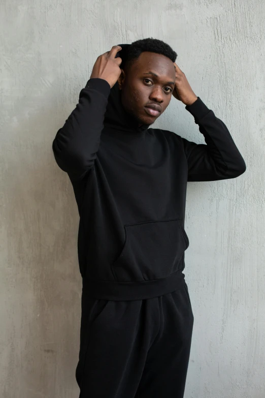 a man standing against a wall with his hands on his head, black turtle neck shirt, wearing a hoodie and sweatpants, thumbnail, black main color