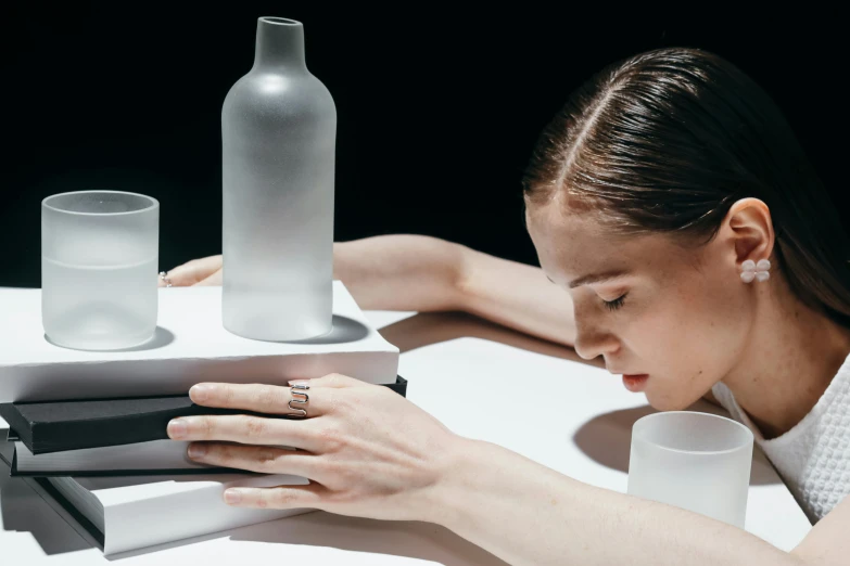 a woman sitting at a table with a bottle and a stack of books, inspired by Anna Füssli, minimalism, porcelain highlighted skin, issey miyake, glassware, award - winning