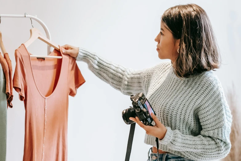 a woman holding a camera in front of a rack of clothes, profile pic, product design shot, inspect in inventory image, pokimane