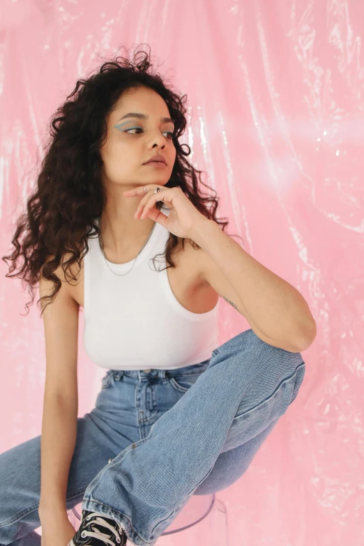 a woman sitting on a stool in front of a pink background, inspired by Elsa Bleda, trending on pexels, renaissance, outfit : jeans and white vest, curly haired, wearing : tanktop, on a white table