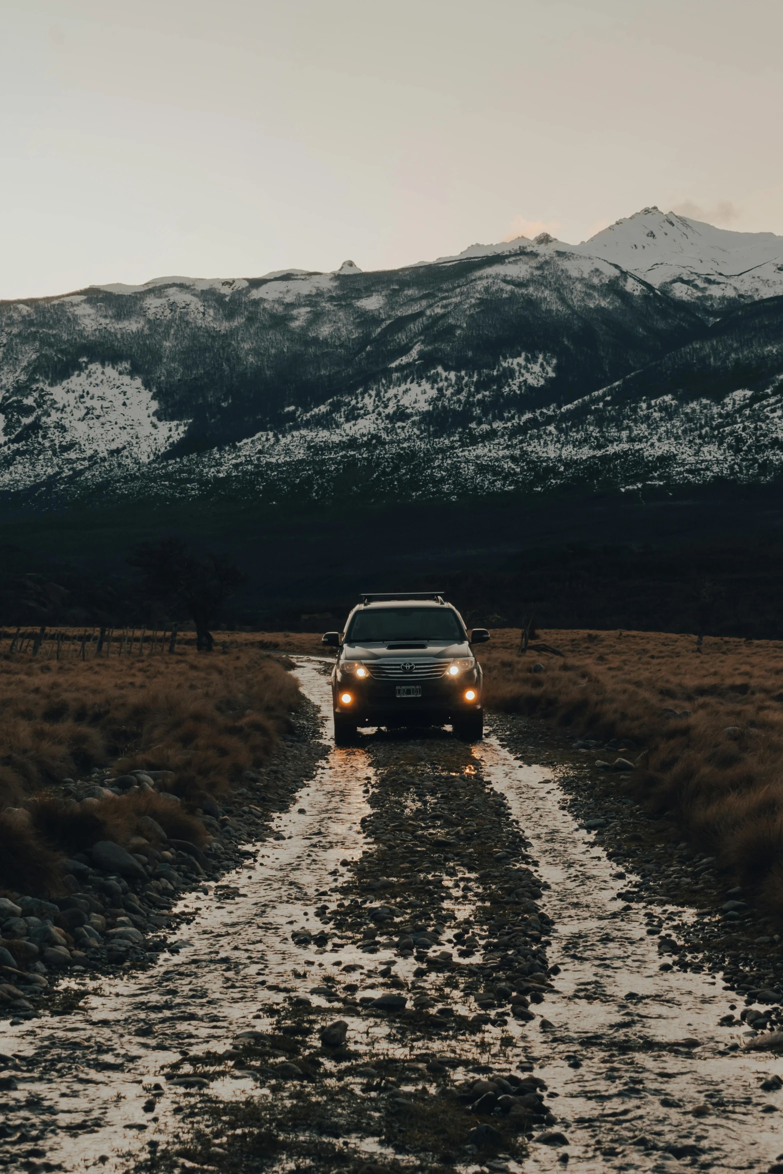 a car driving down a dirt road with mountains in the background, a picture, unsplash contest winner, evening lights, winter, dry river bed, new zealand