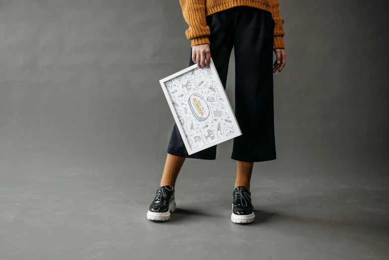 a woman in a yellow sweater holding a picture, inspired by Eden Box, graffiti, white finish, on grey background, carrying a tray, for displaying recipes