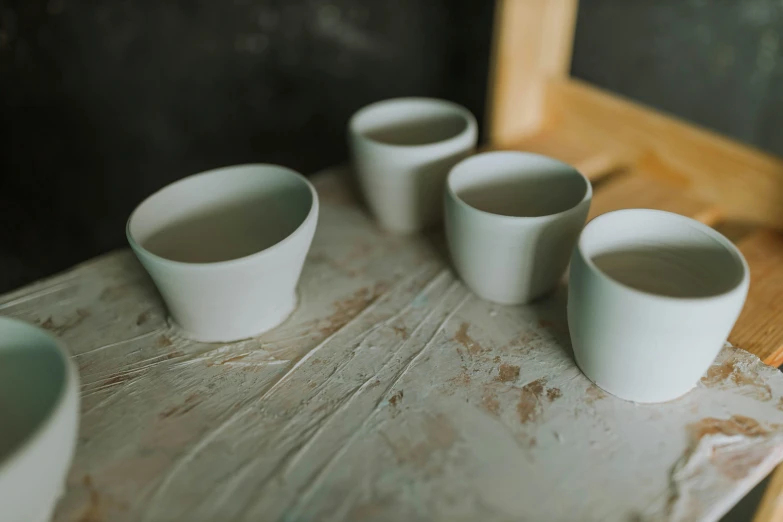 a group of cups sitting on top of a wooden table, a still life, inspired by Agnes Martin, featured on unsplash, process art, white clay, four, middle shot waist up, cast