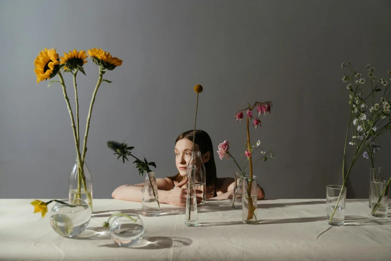 a woman sitting at a table with flowers in vases, a still life, unsplash, floating bodies, ignant, tall, glassware