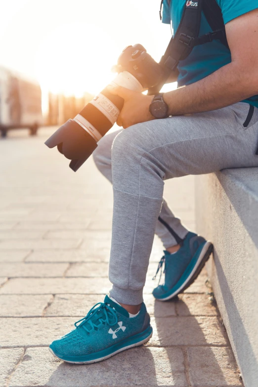 a man sitting on a ledge holding a camera, wearing fitness gear, legs intertwined, on sidewalk, vibrant light