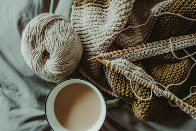 a cup of coffee sitting on top of a bed next to a knitted blanket, trending on pexels, arts and crafts movement, yarn, green and brown clothes, ivory, stitching