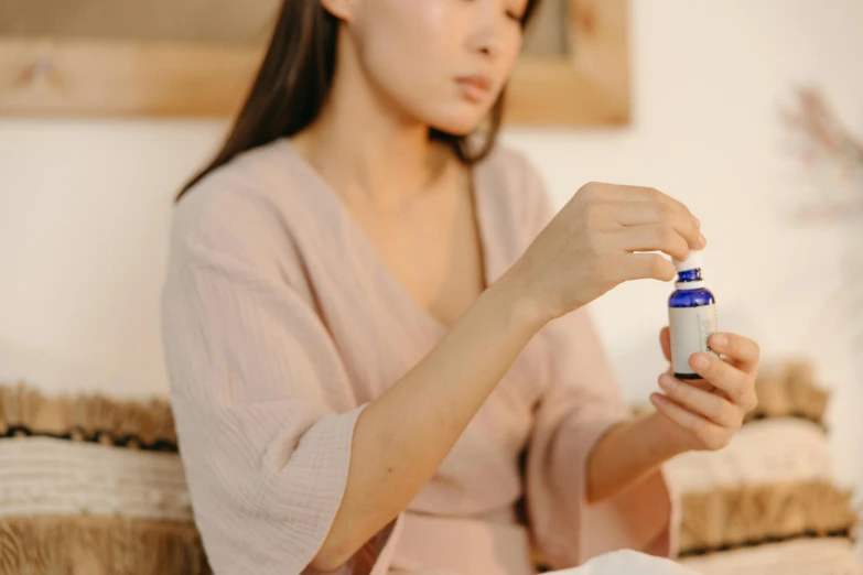a woman sitting on a couch holding a bottle of liquid, a picture, by Emma Andijewska, trending on pexels, apothecary, japanese collection product, wearing a blue robe, glue dropping