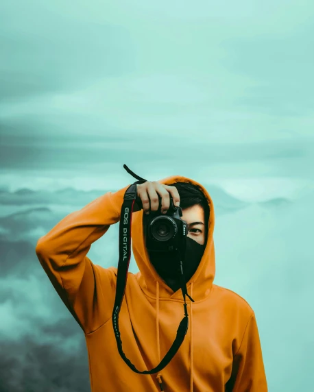 a person taking a picture with a camera, inspired by Elsa Bleda, pexels contest winner, orange balaclava, covered in clouds, non-binary, green hood