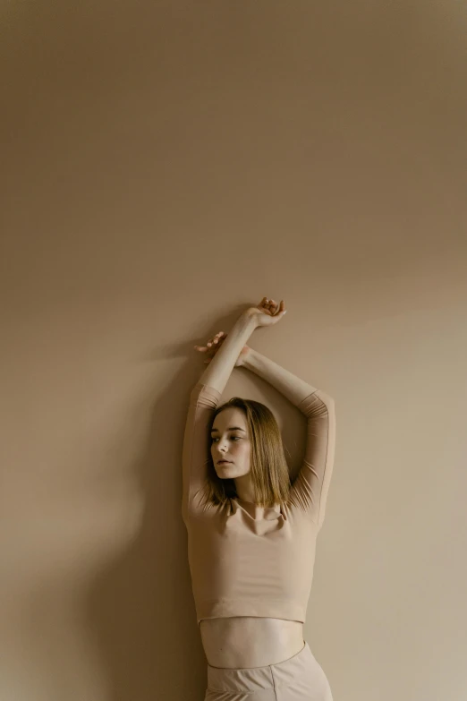 a woman standing against a wall with her arms in the air, inspired by Elsa Bleda, arabesque, saoirse ronan, sand - colored walls, plain background, ignant