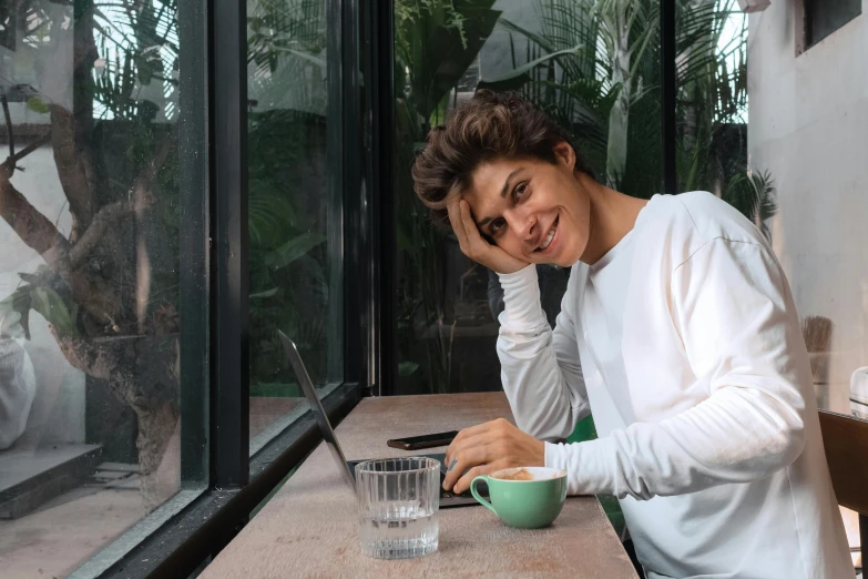 a woman sitting at a table in front of a laptop, inspired by Luca Zontini, happening, androgynous male, relaxing and smiling at camera, diego fernandez, sitting in a cafe alone