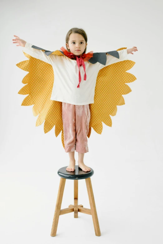 a little girl that is standing on a stool, inspired by Elsa Beskow, unsplash, dada, with real wings, orange and yellow costume, cardboard, full product shot