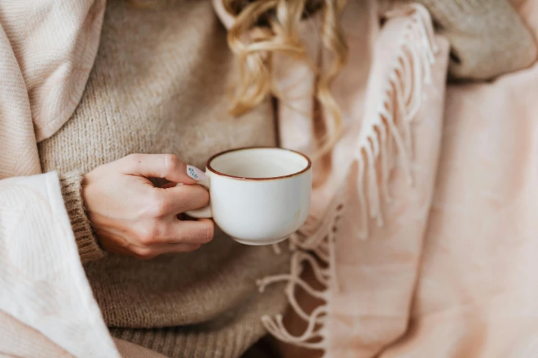 a woman sitting on a bed holding a cup of coffee, by Emma Andijewska, trending on pexels, ivory and copper, wearing a white sweater, pink, wearing a scarf