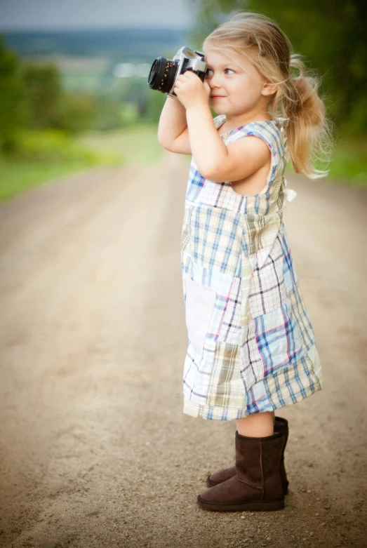 a little girl taking a picture with a camera, inspired by Dorothea Lange, symbolism, cute checkerboard sundress, knee-high boots, dirt road, full color photograph