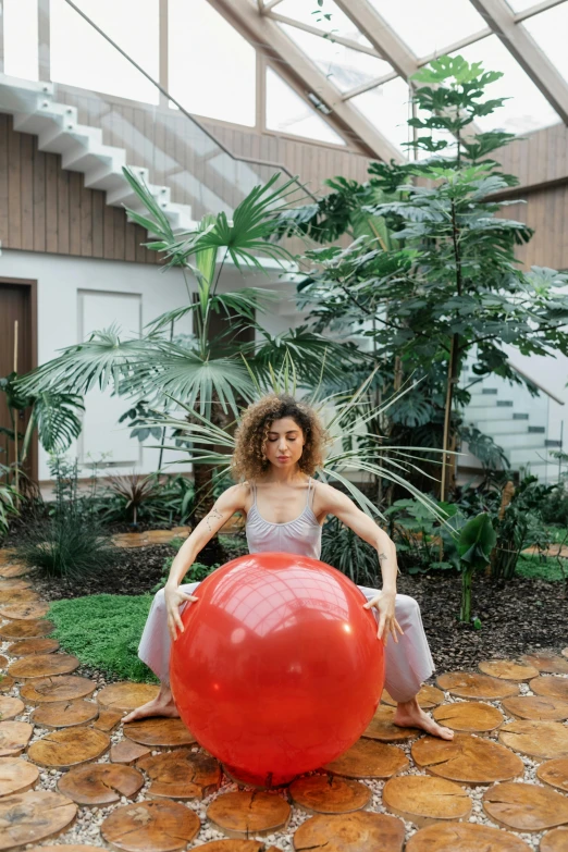 a woman sitting on top of a large red ball, with palm trees in the back, atrium, portrait featured on unsplash, doja cat