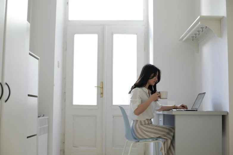 a woman sitting at a desk using a laptop computer, pexels contest winner, minimalism, in doors, muted palette mostly white, panoramic view of girl, eating