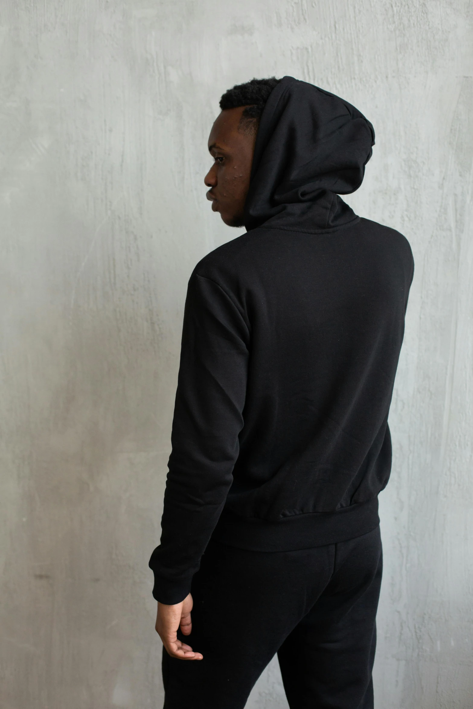 a man standing in front of a wall wearing a black hoodie, by Nina Hamnett, back view also, wearing a track suit, profile image, dark skinned