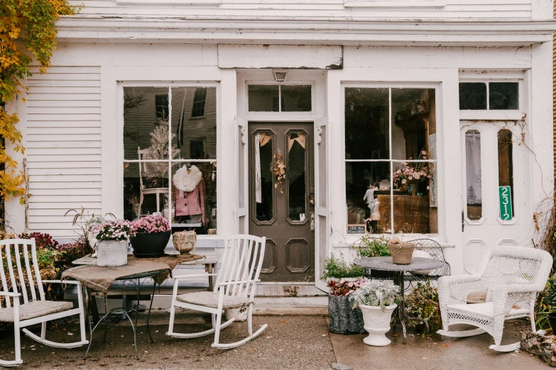 a couple of white rocking chairs sitting in front of a store, by Pamela Drew, trending on unsplash, folk art, pink door, new england architecture, dwell, more and more flowers