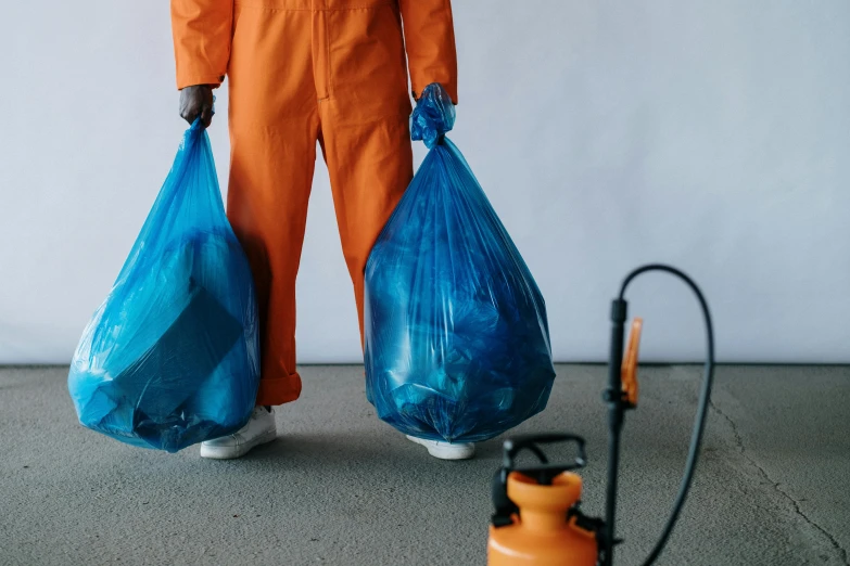 a man in an orange coverall holding two blue bags, by Julia Pishtar, pexels contest winner, plasticien, sweeping, black mold, 4l, diaper disposal robot