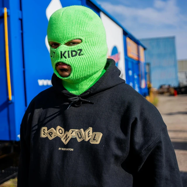 a man wearing a ski mask standing in front of a train, inspired by Zhu Da, graffiti, wearing green clothing, krzysztof porchowski jr, outlive streetwear collection, kids