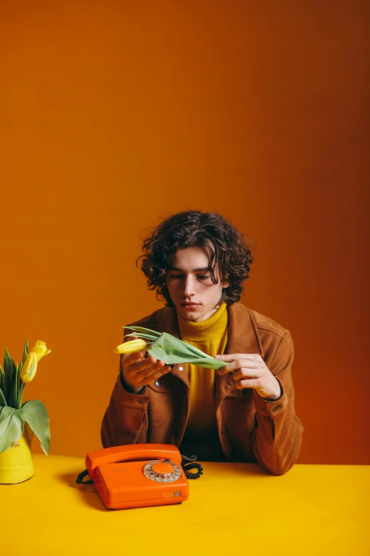 a man sitting at a table with a phone in front of him, an album cover, by artist, trending on pexels, carrying flowers, orange skin, timothee chalamet, colors: yellow