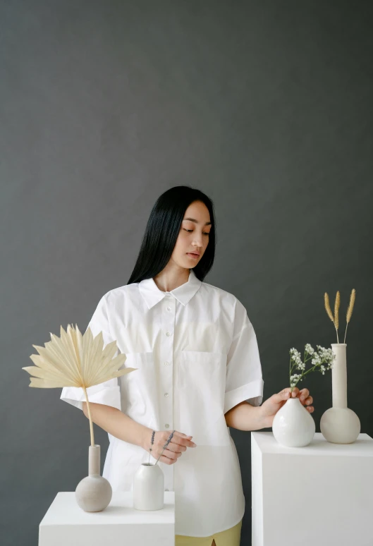 a woman standing in front of some white vases, an album cover, inspired by Kim Tschang Yeul, trending on unsplash, wearing a linen shirt, jakarta, short sleeves, ikebana