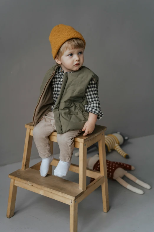 a little boy sitting on top of a wooden step stool, by Nina Hamnett, wearing a vest, olive, beanie, thumbnail