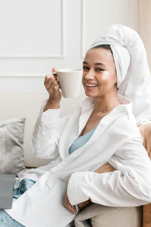 a woman sitting on a couch holding a cup of coffee, wearing white silk robe, skincare, cloth head wrap, wearing a grey robe