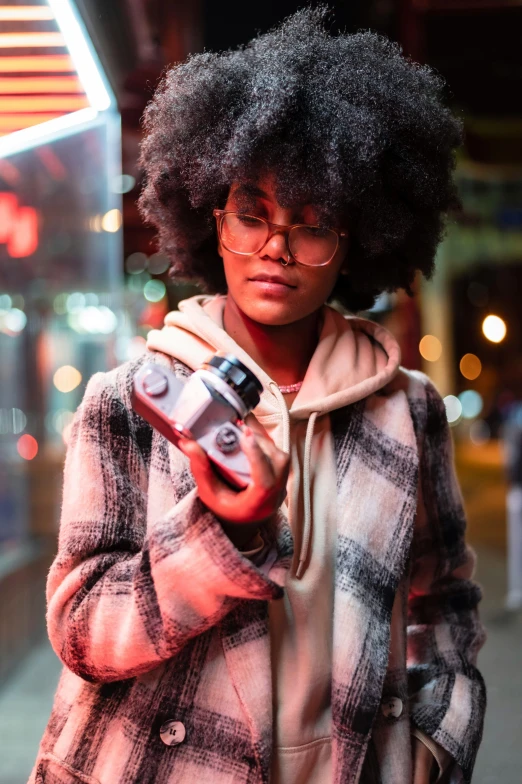 a woman standing on a sidewalk holding a camera, a picture, inspired by Gordon Parks, trending on pexels, afrofuturism, holding flask in hand, vibrant lights, girl wearing hoodie, with square glasses