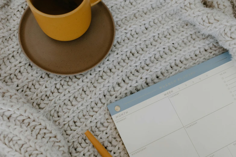 a cup of coffee sitting on top of a desk next to a calendar, a cross stitch, inspired by Agnes Martin, trending on pexels, background image, plush leather pad, grey, knitted mesh material