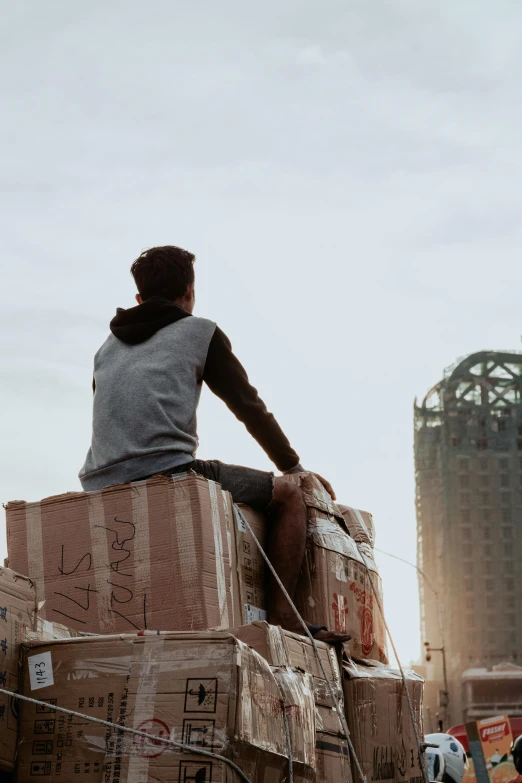 a man sitting on top of a pile of boxes, by Niko Henrichon, pexels contest winner, tall buildings in background, facing away, teenage boy, delivering parsel box