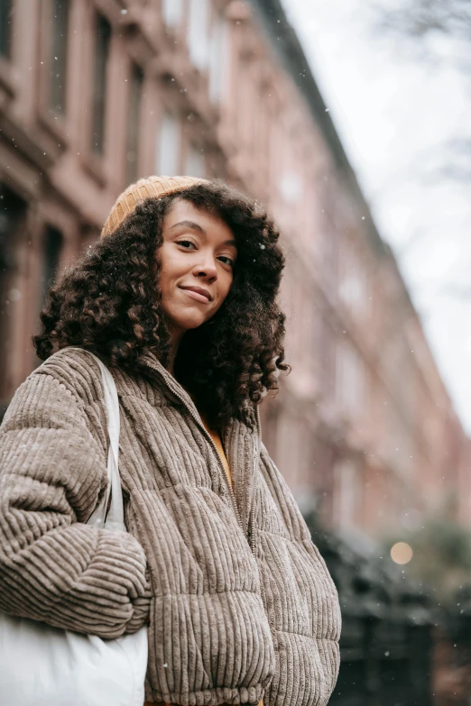 a woman standing on a city street in the snow, trending on pexels, renaissance, curly bangs, wearing hay coat, mixed race woman, muted brown