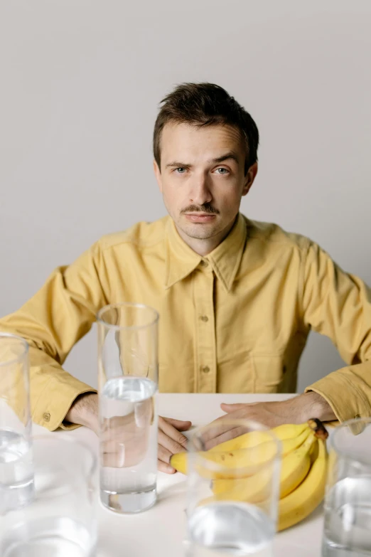 a man sitting at a table with a bunch of bananas, an album cover, by Jessie Alexandra Dick, thin moustache, gold shirt, andrea savchenko, looking straight to camera