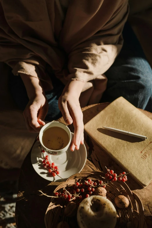 a person sitting at a table with a cup of coffee, a still life, trending on pexels, romanticism, her hands are red roots, “berries, tea drinking and paper lanterns, dreamy feeling