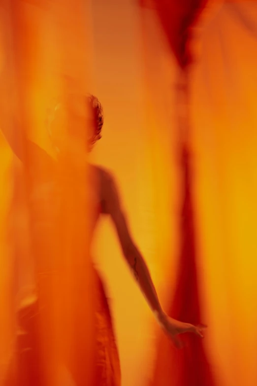 a blurry image of a woman in a dress, figuration libre, light red and orange mood, behind red curtains, nick knight, an orange fire in the background