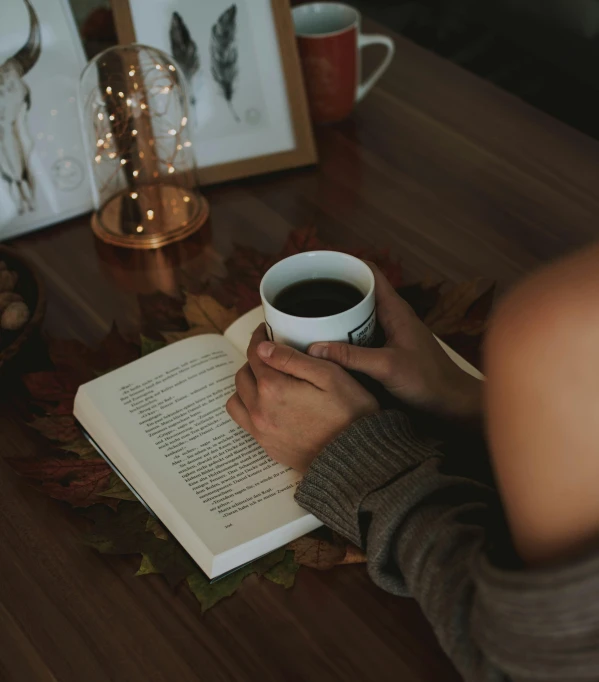 a person sitting at a table with a book and a cup of coffee, 🍂 cute, over-shoulder shot, brown, glowing