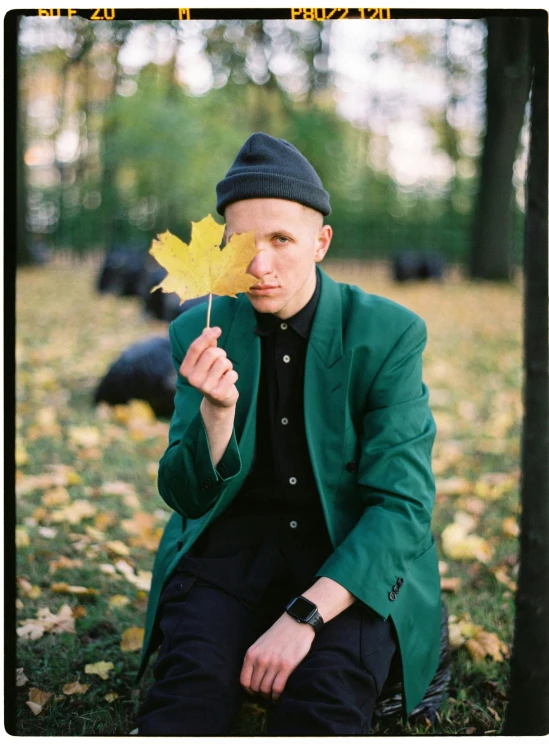 a man sitting on the ground holding a leaf, an album cover, by Attila Meszlenyi, wearing green jacket, no eyebrows, anna nikonova aka newmilky, autum