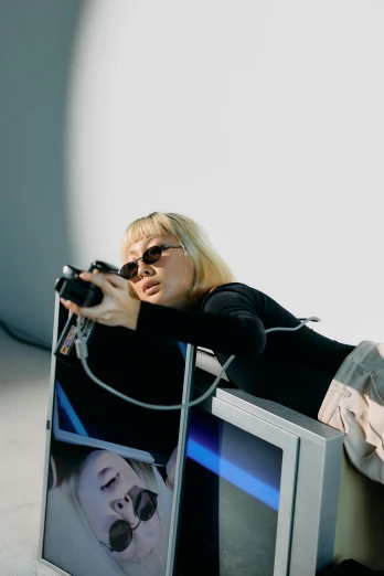 a woman laying on top of a tv while holding a camera, video art, futuristic sunglasses, a blond, in a photo studio, in style of kar wai wong
