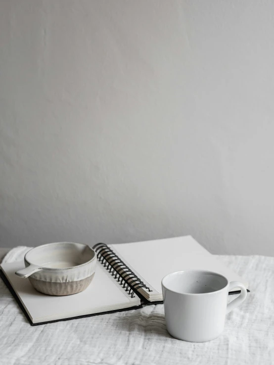 a cup of coffee and a notebook on a bed, a still life, inspired by Agnes Martin, trending on unsplash, minimalism, on kitchen table, organic ceramic white, silver，ivory, high quality product image”