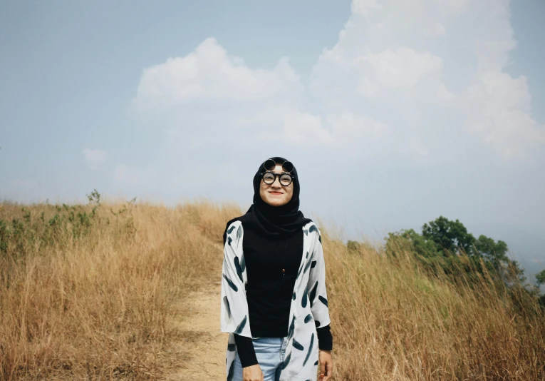 a woman standing in the middle of a field, unsplash, hurufiyya, wearing small round glasses, malaysian, on a hill, wearing a scarf