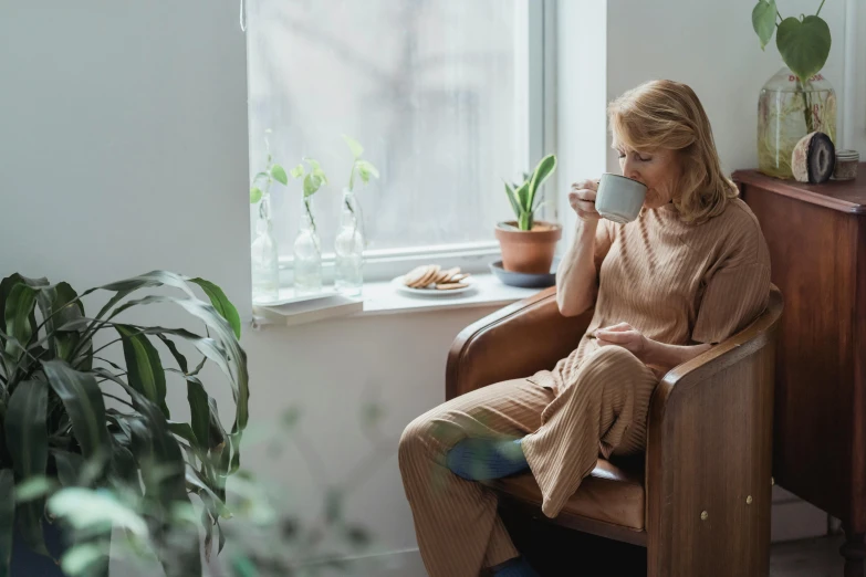 a woman sitting in a chair next to a window, trending on pexels, drinking tea, muted brown, biophilia mood, sitting on chair