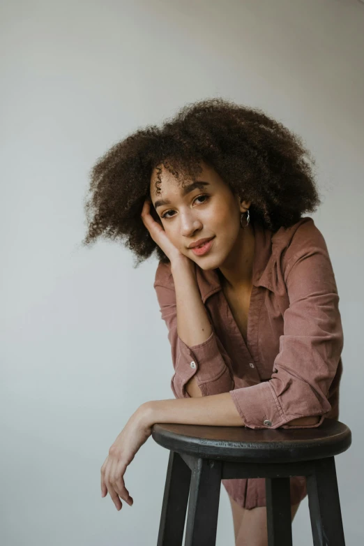 a woman sitting on top of a black stool, a character portrait, trending on pexels, ashteroth, brown fluffy hair, resting head on hands, confident expression