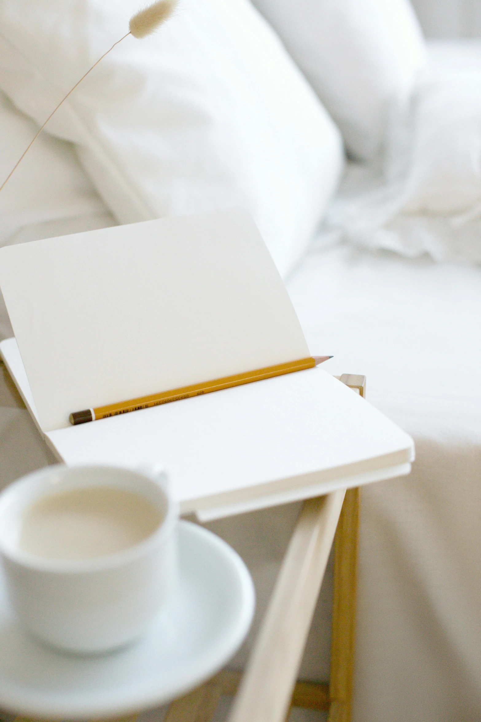 a table with a cup of coffee and a notebook on it, sitting on edge of bed, white with gold accents, holding pencil, promo image