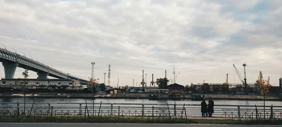 a couple of people standing next to a body of water, a picture, inspired by Elsa Bleda, pexels contest winner, visual art, industrial complex, seen from a distance, rostov city, nice slight overcast weather