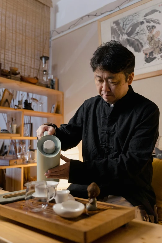 a man pouring tea into a cup on a table, inspired by Kanō Naizen, in a workshop, ryuichi sakamoto, holding flask in hand, ginko showing a new mushi