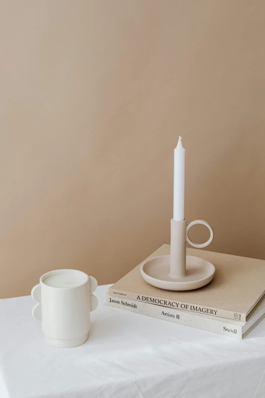 a white candle sitting on top of a table next to a stack of books, an abstract sculpture, inspired by Giorgio Morandi, minimalism, hook as ring, with a white mug, lumi, on simple background