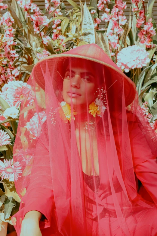 a woman sitting in front of a bunch of flowers, an album cover, inspired by Cecil Beaton, trending on pexels, renaissance, red hat, imaan hammam, veil, pink and yellow