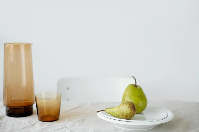 a white plate topped with a pear next to a glass, a still life, unsplash, minimalism, light brown, john pawson, background image, amber