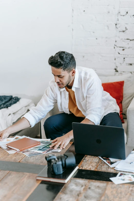 a man sitting on a couch working on a laptop, by Julia Pishtar, trending on pexels, visual art, papers on table, wearing business casual dress, lots of pictures, fashion designer