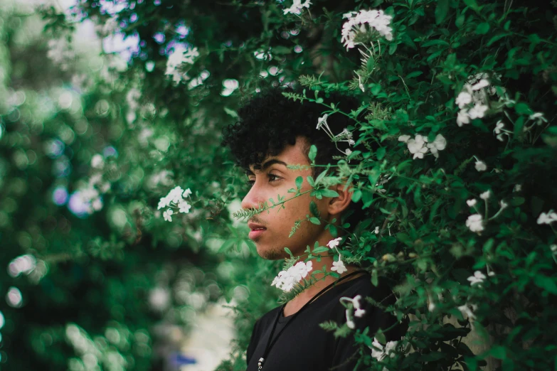 a man standing in front of a bush with white flowers, pexels contest winner, black teenage boy, middle eastern skin, profile image, green and black colors