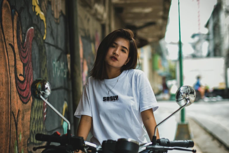 a woman sitting on a motorcycle in front of a graffiti covered wall, inspired by Rudy Siswanto, unsplash contest winner, white tshirt, simple logo, smirking, \'obey\'
